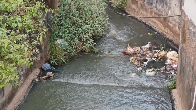 Belagavi, Karnataka, India - December 3, 2025: Polluted urban canal with flowing water, plastic waste and overgrown vegetation showing environmental impact and city drainage conditions.