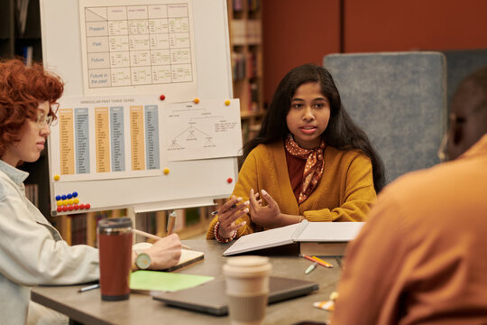 Teenage Asian girl explaining grammar rules to diverse group of teenagers in classroom setting, gesturing with hands while sitting at table with open books and educational charts