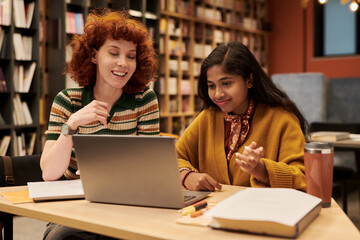 Young woman and Asian teenage girl sitting at table working together on laptop in library, both smiling and engaging in collaborative learning with books nearby