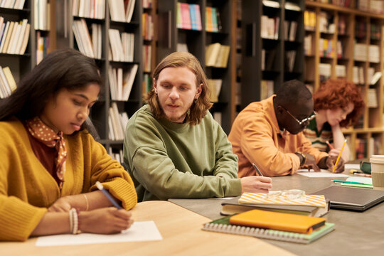 Group of multiethnic teenagers studying together in library, teenage boy talking to Asian teenage girl while Black teenage boy and teenage girl writing notes