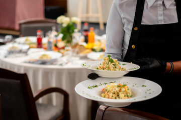 A waiter in a black apron and gloves serves two plates of a fresh, colorful vegetable salad in a restaurant setting with a white tablecloth table set in the background