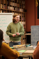 Young man standing in front of flip chart teaching group of people in classroom, holding marker and...