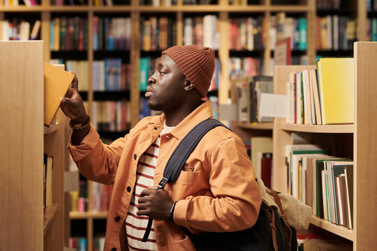 Young Black man browsing bookshelf in library, reaching for book with right hand while carrying backpack over shoulder, standing among shelves filled with books