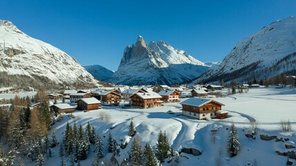 Aerial view of a charming alpine village nestled in a snowy valley with traditional wooden chalets under a bright blue sky and dramatic mountain peak in the background