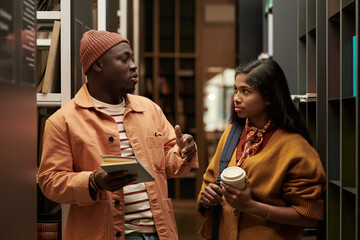 Black young man holding books and gesturing while talking to Asian young woman holding coffee cup, both standing between bookshelves in library or bookstore