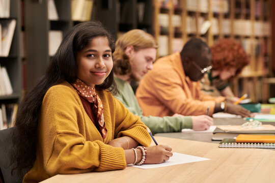 Portrait of teenage Asian girl smiling at camera while sitting at desk in library, multiethnic group of teenagers studying and writing in background, bookshelves