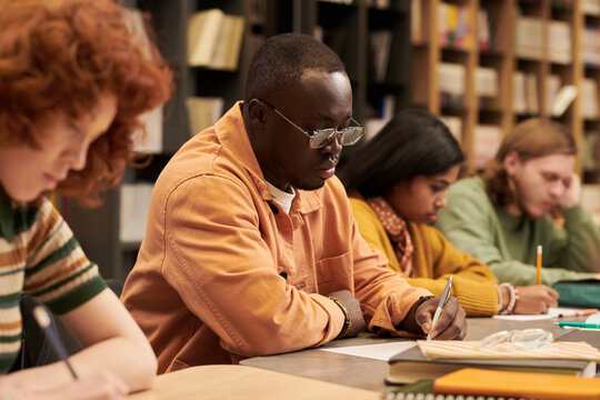 Group of multiethnic teenagers sitting at table writing in notebooks in library, focused on studying and taking notes, bookshelves in background, diverse students in learning