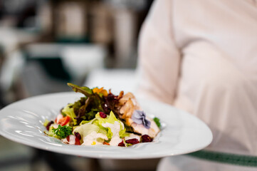 A close-up, selective focus shot of a chef holding a white plate of gourmet chicken salad with mixed greens, dried fruits, and edible flowers.