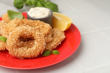 Deep fried squid rings with basil, sauce and slice of lemon on white tiled table, closeup. Space for text