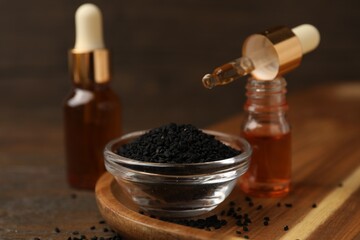 Black cumin seeds in bowl and bottles of essential oil on wooden table, closeup