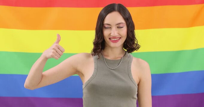 Smiling woman stands against rainbow pride flag showing thumbs up gesture. Supporter person celebrates equality message while enjoys tolerant vibe
