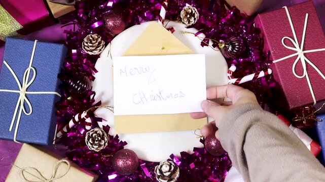Colorful holiday scene featuring wrapped gifts, hand putting a parchment note saying Merry Christmas on a marble stone, pinecones, ribbon, and glittery ornaments 