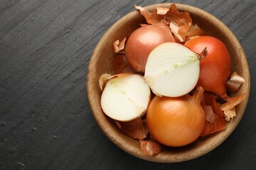 Whole and cut onion bulbs with peels in bowl on black textured table, top view. Space for text