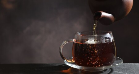 Pouring hot tea into cup from teapot at black table, closeup. Space for text