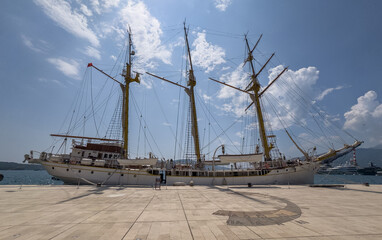 Three-masted tall ship moored at sunlit Tivat Marina, its rigging etched against a blue sky beside a compass-rose plaza.