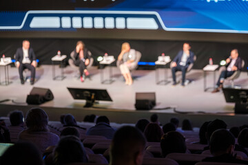 Panel discussion at a conference with five speakers on stage, audience visible in foreground, professional atmosphere, business event, and engaging talks.