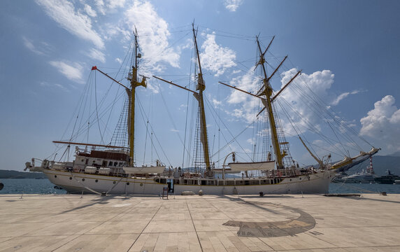 Three-masted tall ship moored at sunlit Tivat Marina, its rigging etched against a blue sky beside a compass-rose plaza.