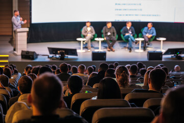 Panel of five speakers on stage during conference talk, audience listening intently in theater setting