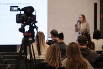 Speaker with microphone presents to attentive audience while camera captures session; large projection screen displays slides; audience seated in rows; professional event atmosphere, 