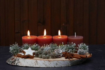 Dritter Advent: Längliches Adventsgesteck mit braunen Kerzen, Zimtsternen, Zapfen, Beeren, Zimtstange und Tannenzweigen vor einer braunen Holzwand mit Textfreiraum.