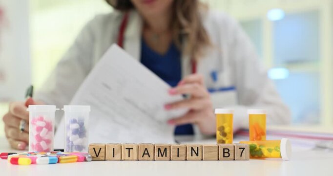 Wooden blocks spell word Vitamin B7 on medical desk. Woman doctor writes notes studying bottles with bright supplement pills scattered around