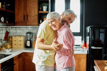 Portrait of happy senior mid aged mature couple prepering meal and having fun dancing and embracing in kitchen at hpme