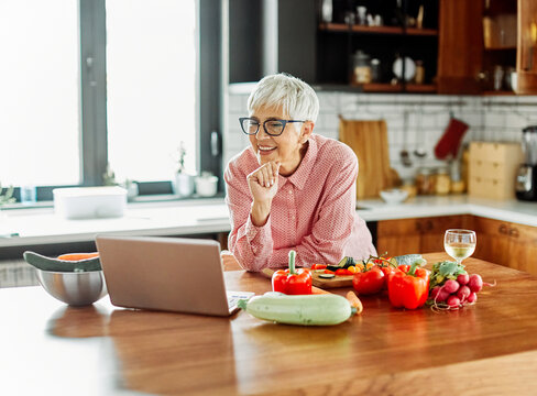 Fototapeta Portrait of happy senior mid aged mature woman prepering meal with fresh vegatebles and following internet instructions for a recipe on a laptop computer  or looking at video  or website app and drink