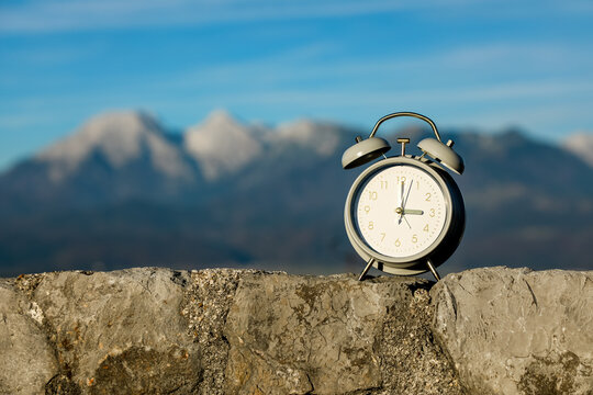 Classic alarm clock resting on a stone wall with Alpine mountains in the background, symbolizing the seasonal shift from Daylight Saving Time to standard winter time as the clock hands are set back.