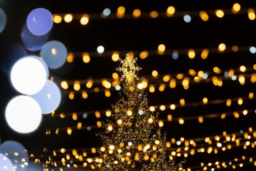 A festive outdoor scene featuring a Christmas tree adorned with golden lights, surrounded by strings of warm, glowing bokeh lights against a dark night sky
