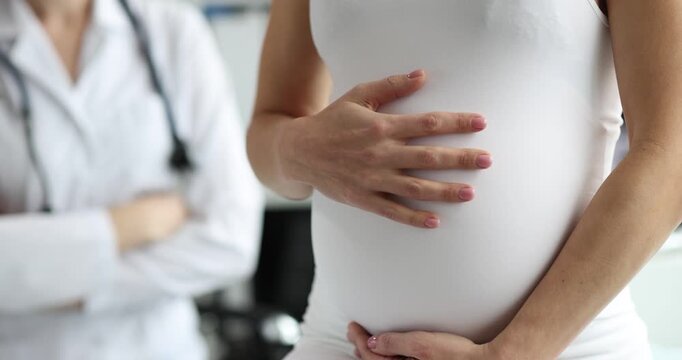 Pregnant woman holds round belly with both hands in clinic. Doctor figure stays hands-crossed behind listening to pregnancy story during appointment