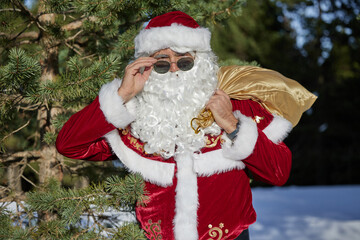 Santa Claus lifting sunglasses while carrying golden gift bag in snowy forest.