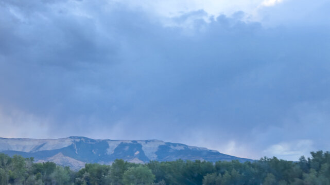 Storm clouds with virga over green treeline and layered mesa at dusk