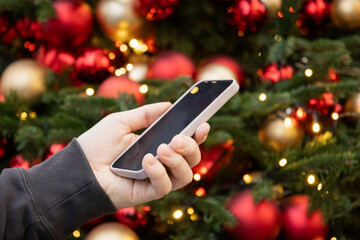 Smartphone in mans hand against Christmas tree with decorative and light garland. Screen display for Festive Xmas Holiday Background. Close up. Christmas advertising, smartphone app template
