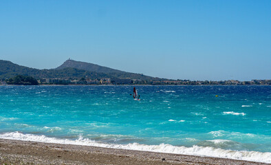 Windy turquoise sea with small waves rolling onto pebble beach, lone windsurfer sailing near distant coastal town and green mountain under clear blue sky.