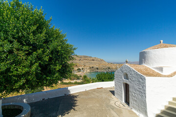 Whitewashed Greek chapel with stone roof and cross overlooking a blue bay and arid hills under clear summer sky in a peaceful Mediterranean coastal landscape.