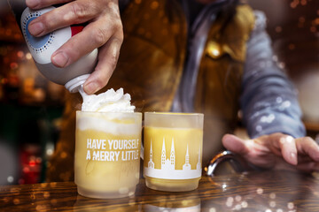 Hand of a waiter spraying whipped cream into glass mugs filled with hot eggnog punch, alcoholic hot...