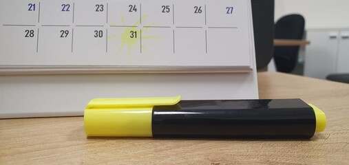A yellow and black highlighter rests on a wooden desk in an office setting. Behind it, a desk calendar displays a yellow highlight around the 31st, suggesting a marked reminder or important date.