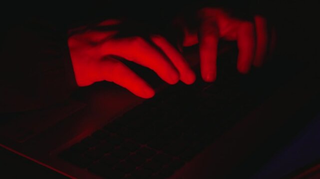 A close-up shot of hands typing on a laptop keyboard, dramatically lit with red light in a dark environment, evoking themes of hacking, cybersecurity threats, or illicit digital activity.