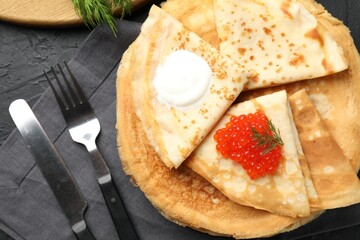 Delicious crepes with red caviar and sour cream served on black textured table, flat lay