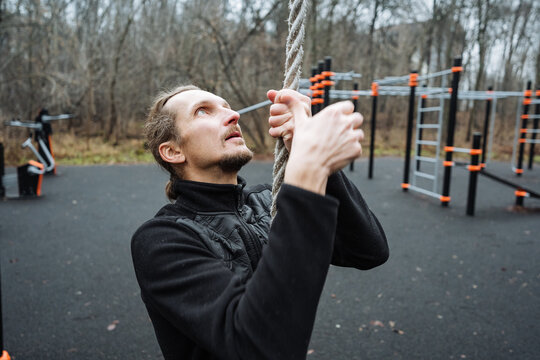 caucasian male practicing obstacle course training, man exercises outdoors on metal bars during autumn daylight, individual using chalked hands to ascend ropes at rustic outdoor fitness area