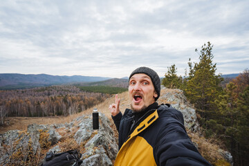 Caucasian blogger reacting to dramatic cliff view with astonished expression, mouth open, hands animated, layered landscape behind, cloudy sky and rugged rocks adding dramatic atmosphere