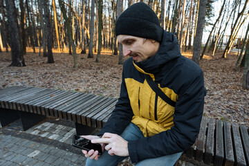 autumn park scene, person sitting on park bench during fall season with phone in hand, man dressed in yellow jacket examining his phone while seated outdoors in fall park setting
