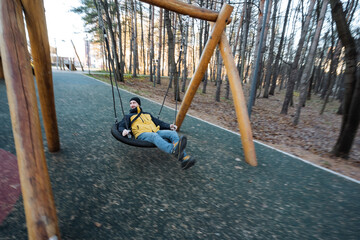gentle sunlight bathes peaceful playground moment, casual male enjoying tranquil park environment during afternoon, serene setting with man resting on swing amid city park surroundings