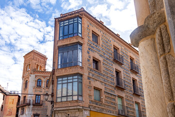 A patterned brick corner building in Toledo with iron balconies and bay windows beside a historic bell tower with arched openings under a blue, cloudy sky