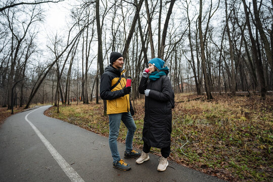 couple walking on trail having conversation, animated gestures and easy pace, winding paved path through bare trees, casual outerwear and mutual attention during autumn stroll