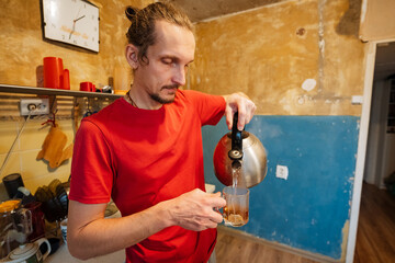 man pouring hot water into cup with kettle, steam rising in compact kitchen, red shirt and calm posture, slow morning ritual and soothing beverage moment in worn apartment interior