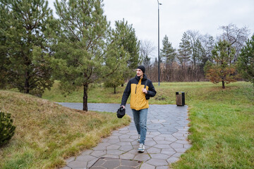 urban traveler walking, individual dressed for travel strolling through green urban park, caucasian male in bright outerwear navigating pathway with travel equipment in outdoor setting