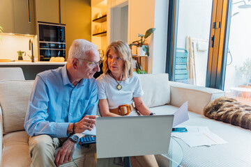 Senior couple calculating household budget with laptop and documents, managing family finances for retirement planning, reviewing bills and expenses at home