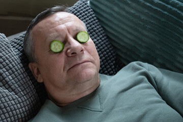 Resting caucasian man lying on sofa with cucumber slices on eyes, practicing simple home skincare and relaxation indoors. Home Leisure Activities