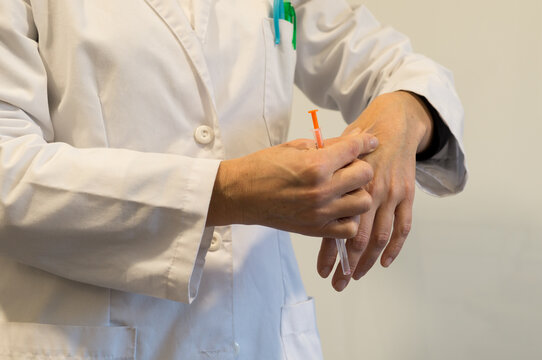 A close-up of a person in a lab coat holding a small syringe and lightly pinching the skin on the back of their hand, preparing for a medical procedure.